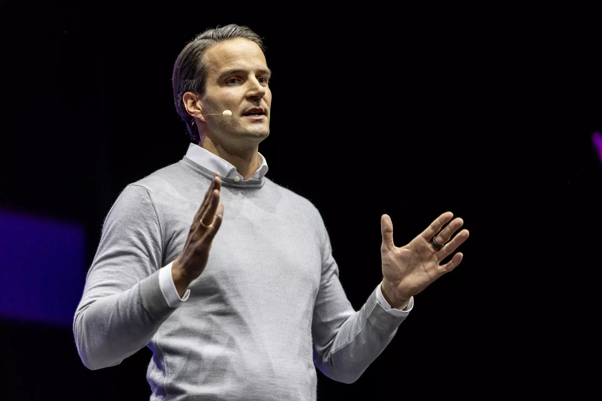 Person im grauen Pullover mit Headset spricht gestikulierend auf Bühne vor dunklem Hintergrund.