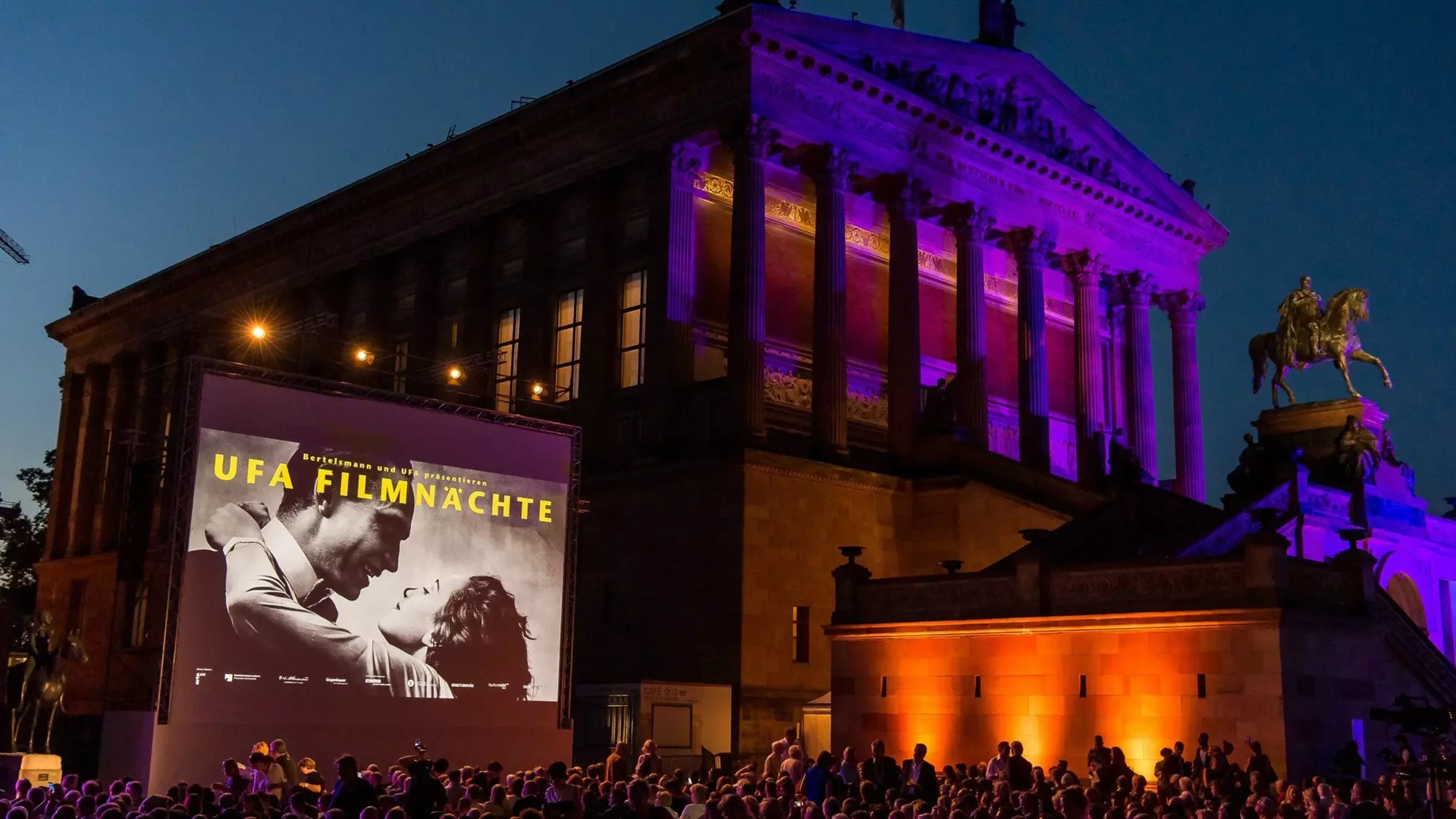 Open-air film screening at night with a large screen set against a monumental building façade. Audience in the foreground and classical architecture with colored lighting in the background.