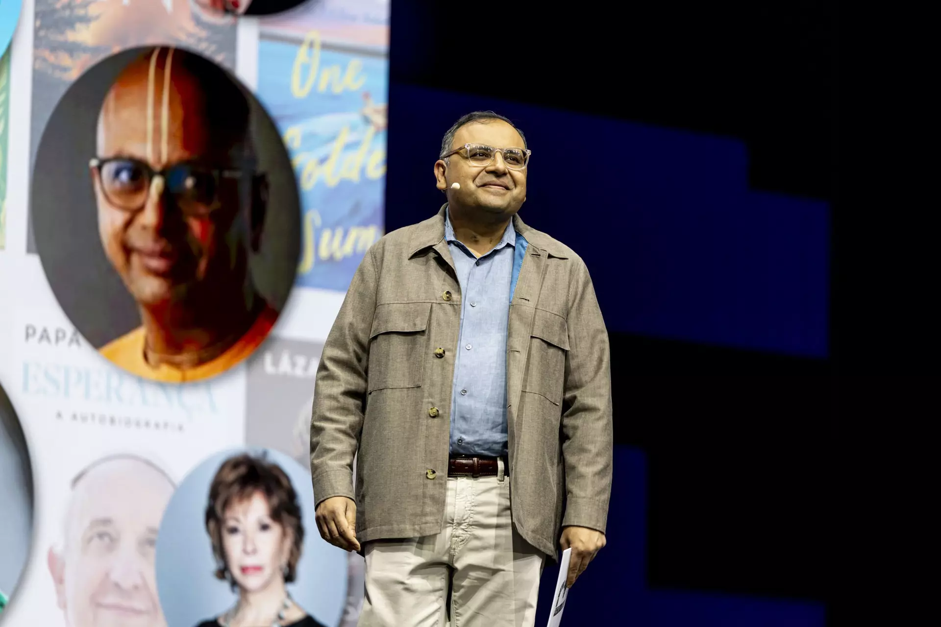Person mit Brille und Jacke steht auf Bühne vor Plakatwand mit Porträtfotos.