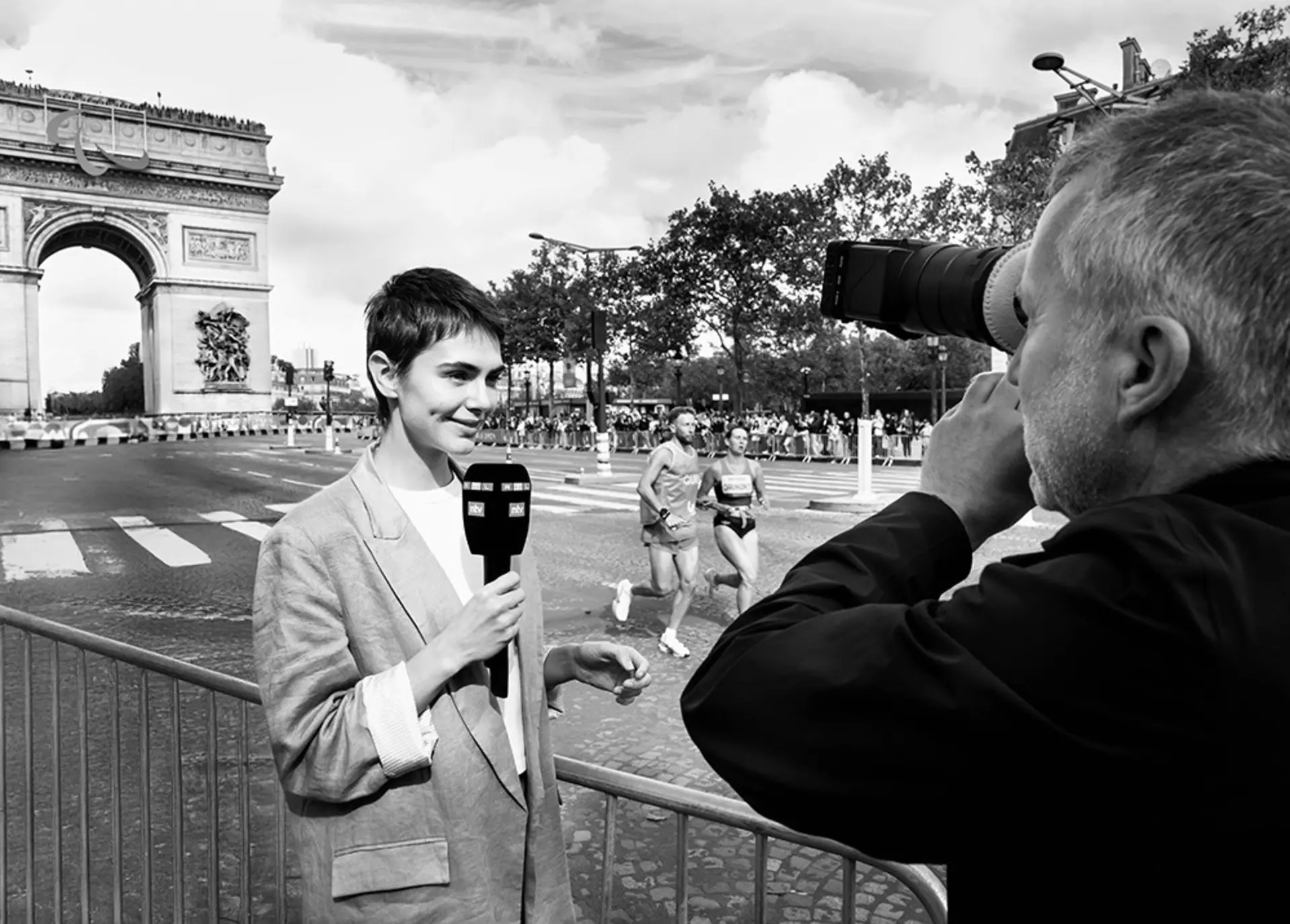 Person mit Mikrofon vor einer Person mit Filmkamera, im Hintergrund zwei Profiläufer in Paris vor dem Arc de Triomphe.