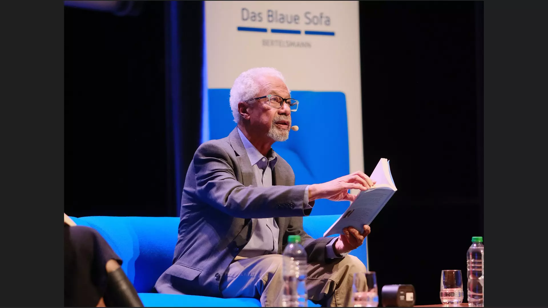 A Black man with white hair and a beard sits on a blue sofa on a stage, holding a large paperback and looking ahead.