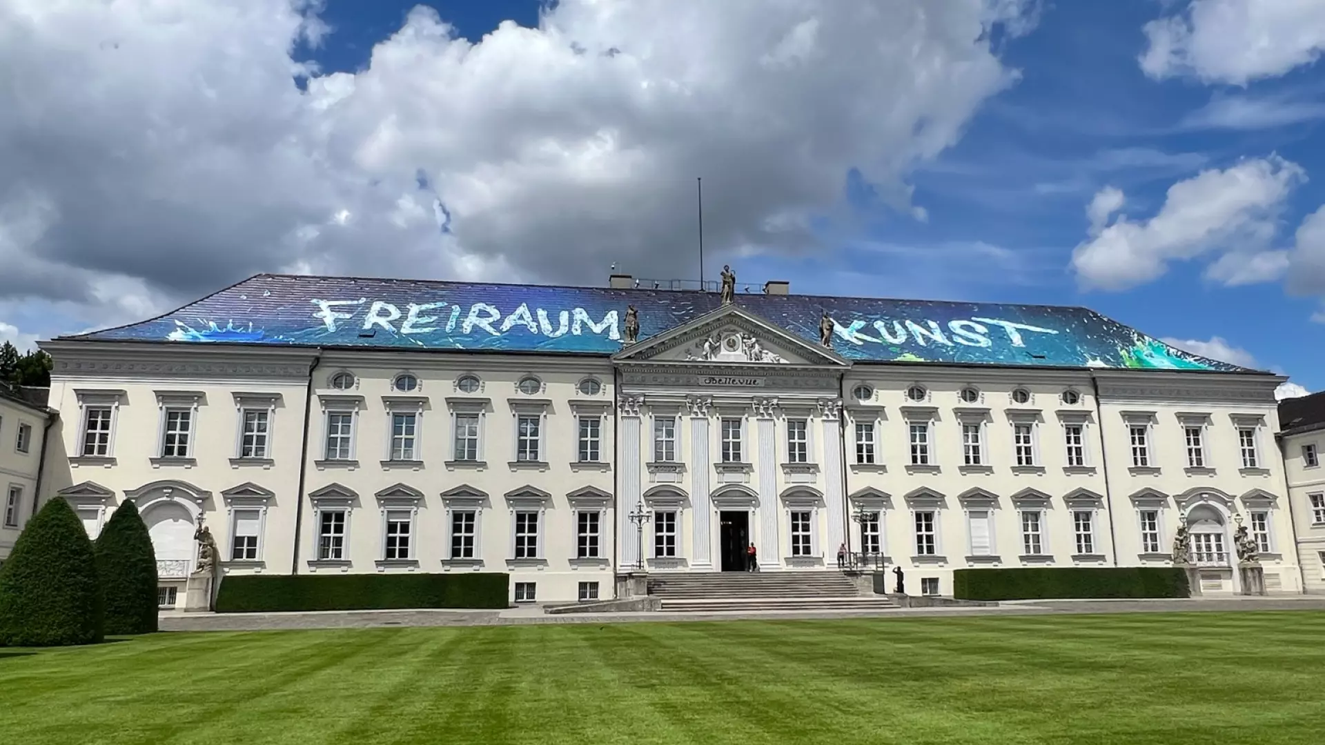 On the roof of Bellevue Palace, you can see the words “Freiraum Kunst.” The palace is bathed in sunlight; you can see the green lawn in front of it and clouds against a blue sky.