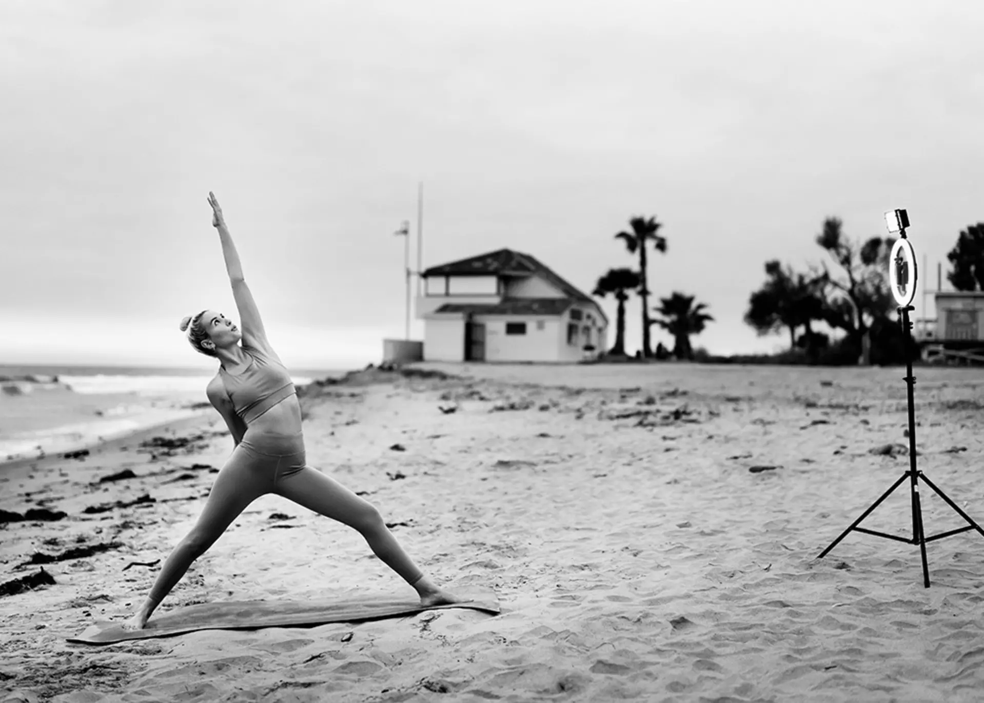 Person macht Yoga-Pose am Strand vor Stativ mit Ringlicht.