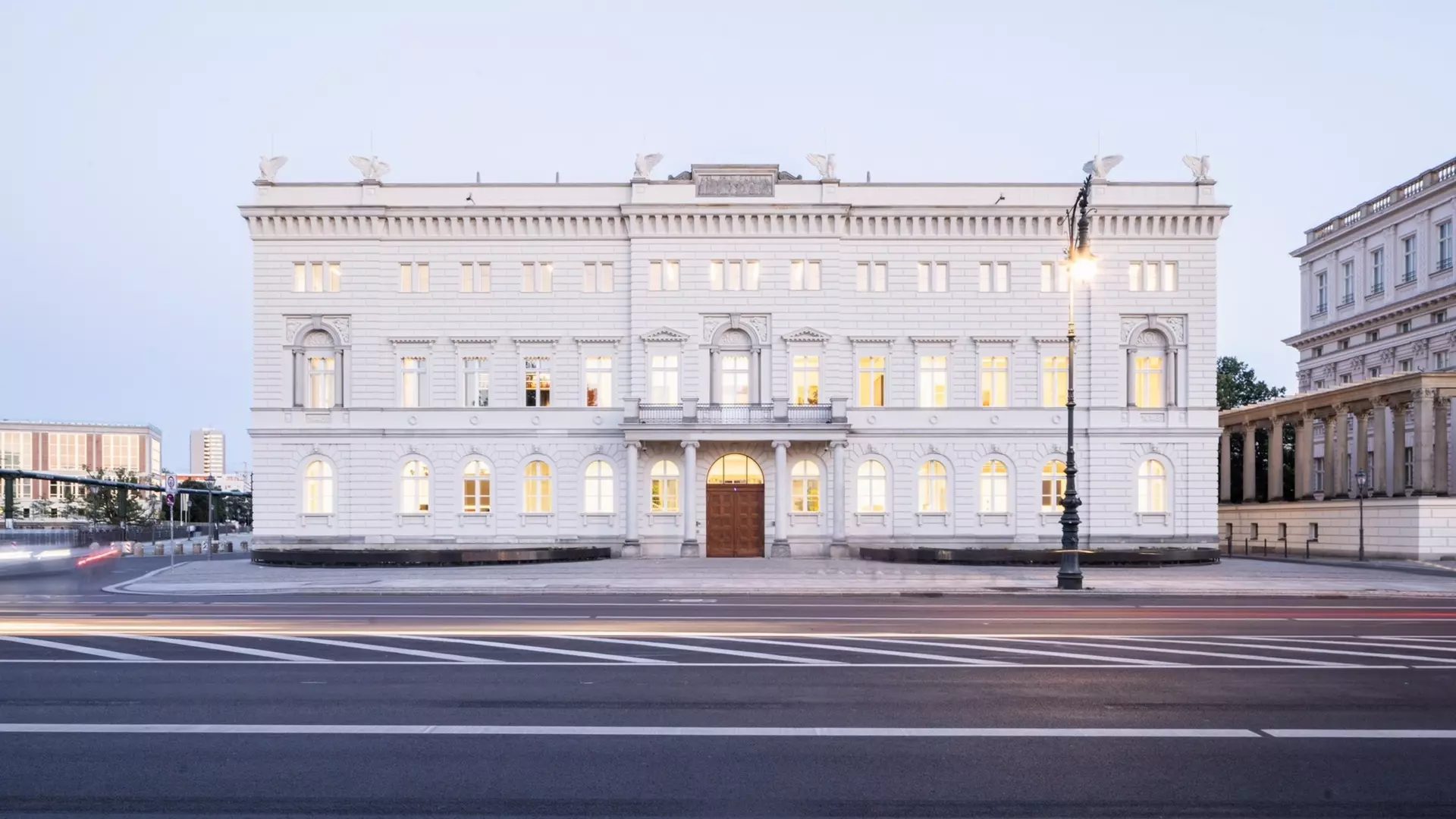 Symmetrische Fassade eines hellen klassizistischen Gebäudes bei Dämmerung, beleuchtete Fenster, Straße im Vordergrund.