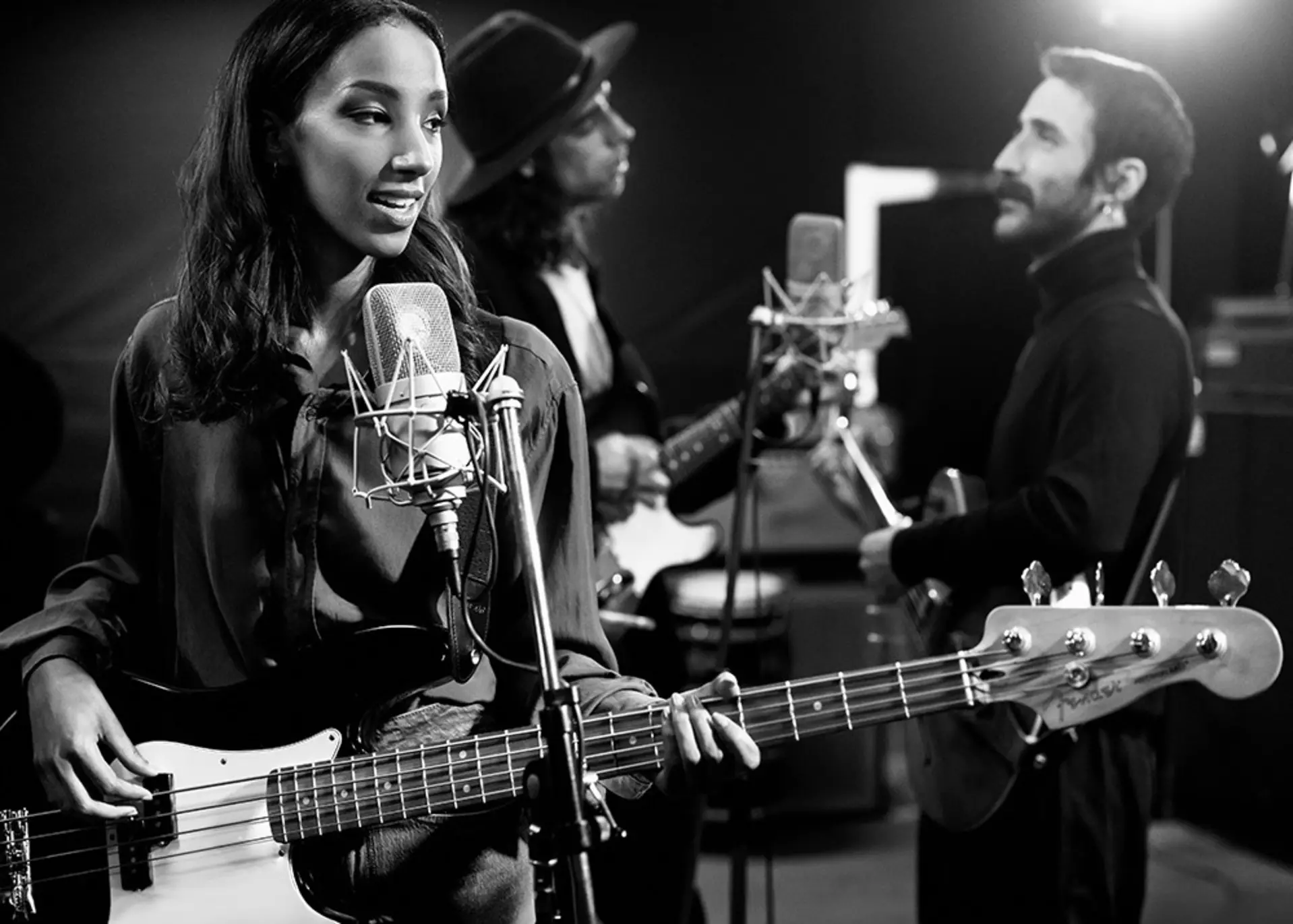 Drei Personen stehen in einem Musikstudio vor Mikrofonen mit E-Gitarren in der Hand.