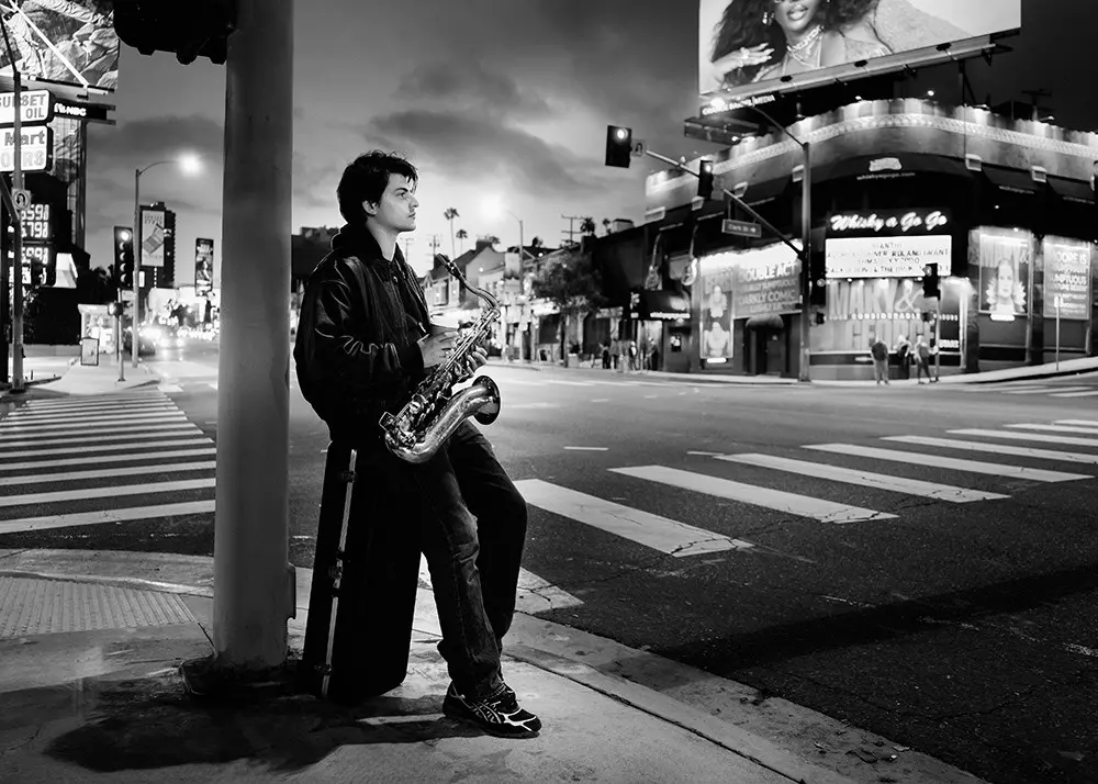 Person mit Saxofon an einer Straßenkreuzung in Hollywood, im Hintergrund beleuchtete Werbetafeln.