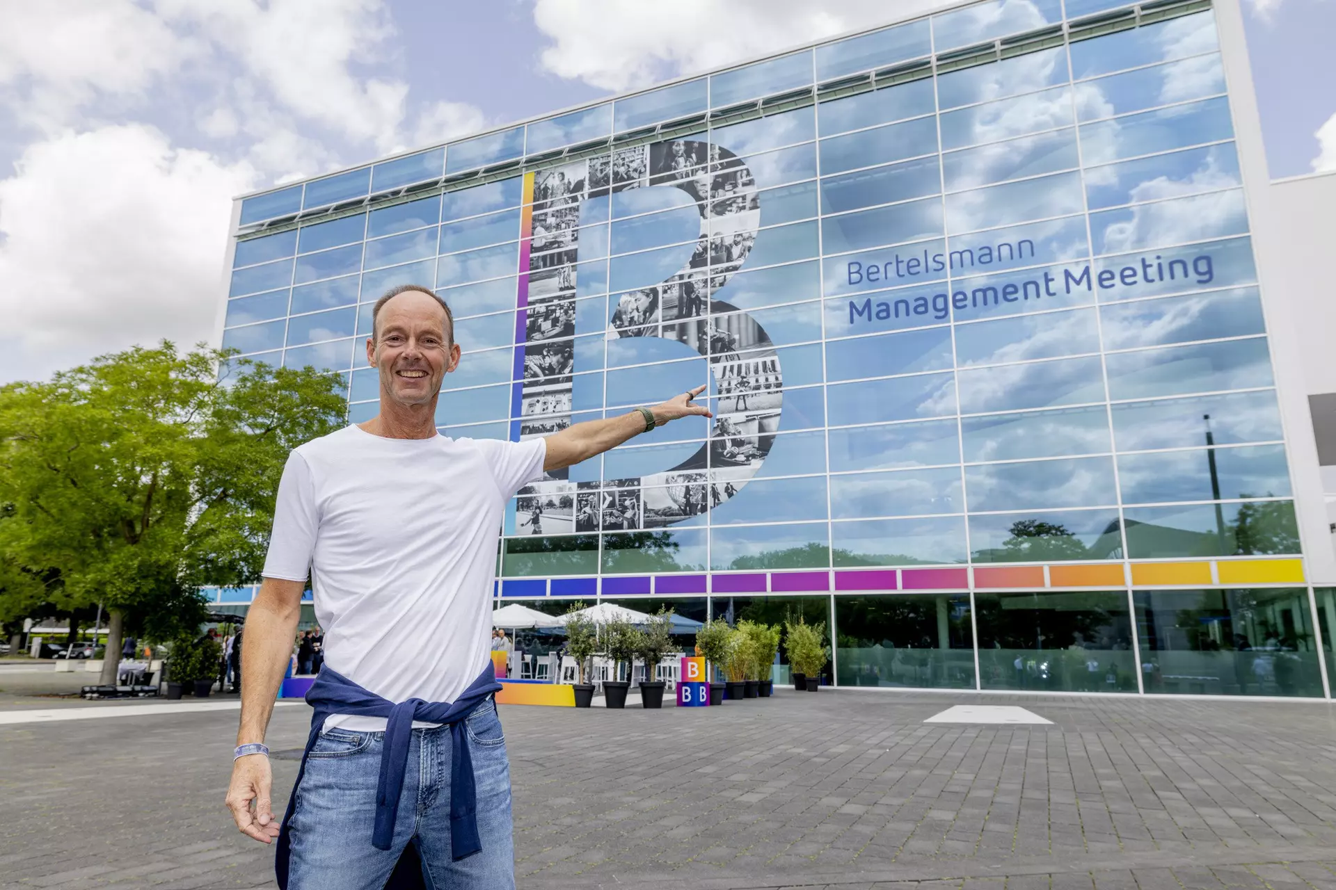 Person im T-Shirt zeigt auf Glasfassade mit großem „B“ und Schriftzug „Bertelsmann Management Meeting“.