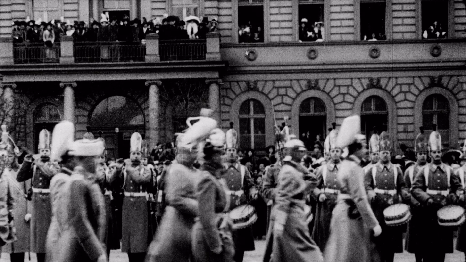 Historische Fotografie mit festlich uniformierten Soldaten, manche mit Trommeln, im Vorbeimarsch vor Gebäudefassade mit Bogenfenstern und einem Balkon voller Zuschauer