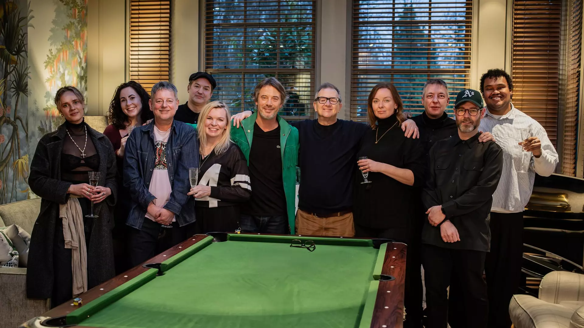 Eleven people stand in front of a billiard table. Some of them are holding champagne flutes in their hands.