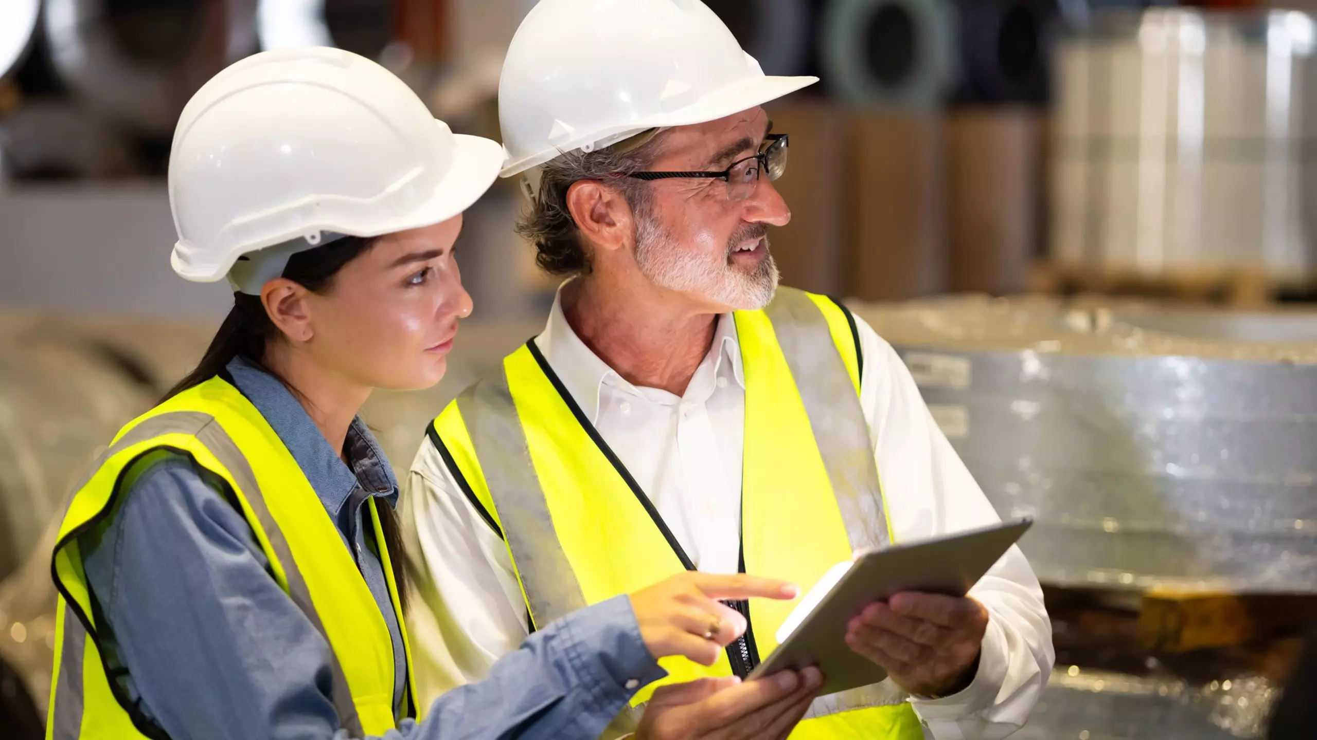Person in orange Schutzkleidung und Helm steht in Industriehalle und nutzt ein Tablet zur Kontrolle von Arbeitsabläufen.
