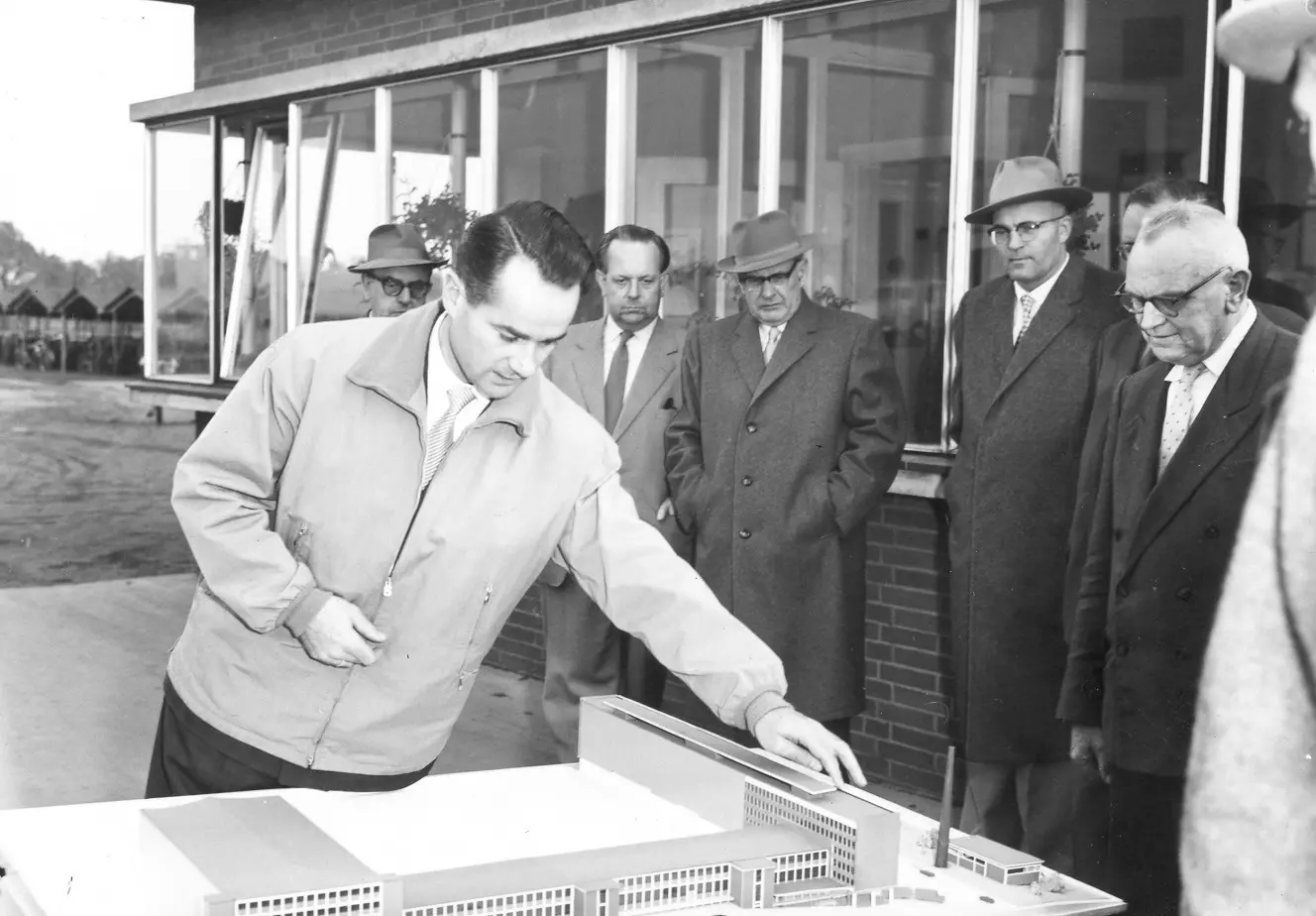 Reinhard Mohn stands in front of a miniature building complex. In the background, other people are looking at the building model.