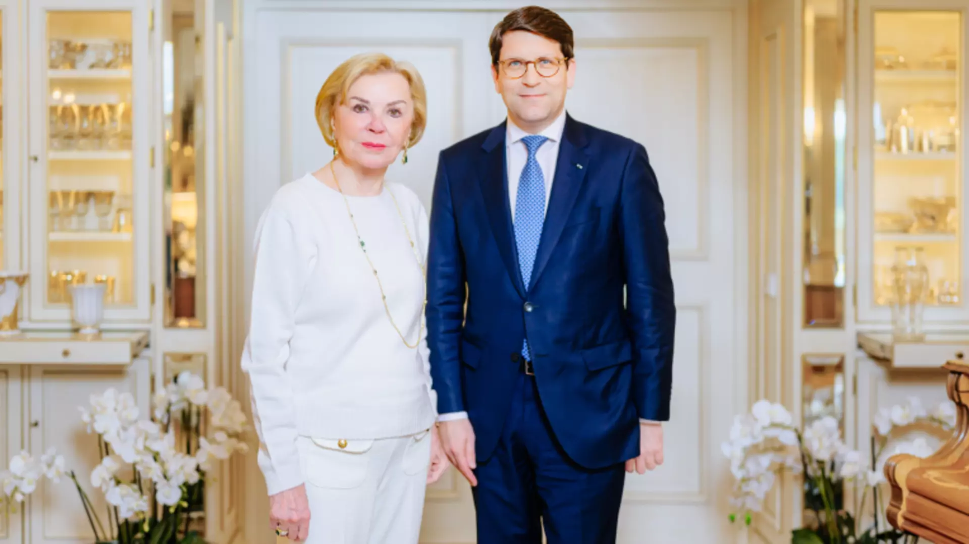 Liz Mohn dressed in white and Alexander Neef in a blue suit and tie stand side by side and look into the camera
