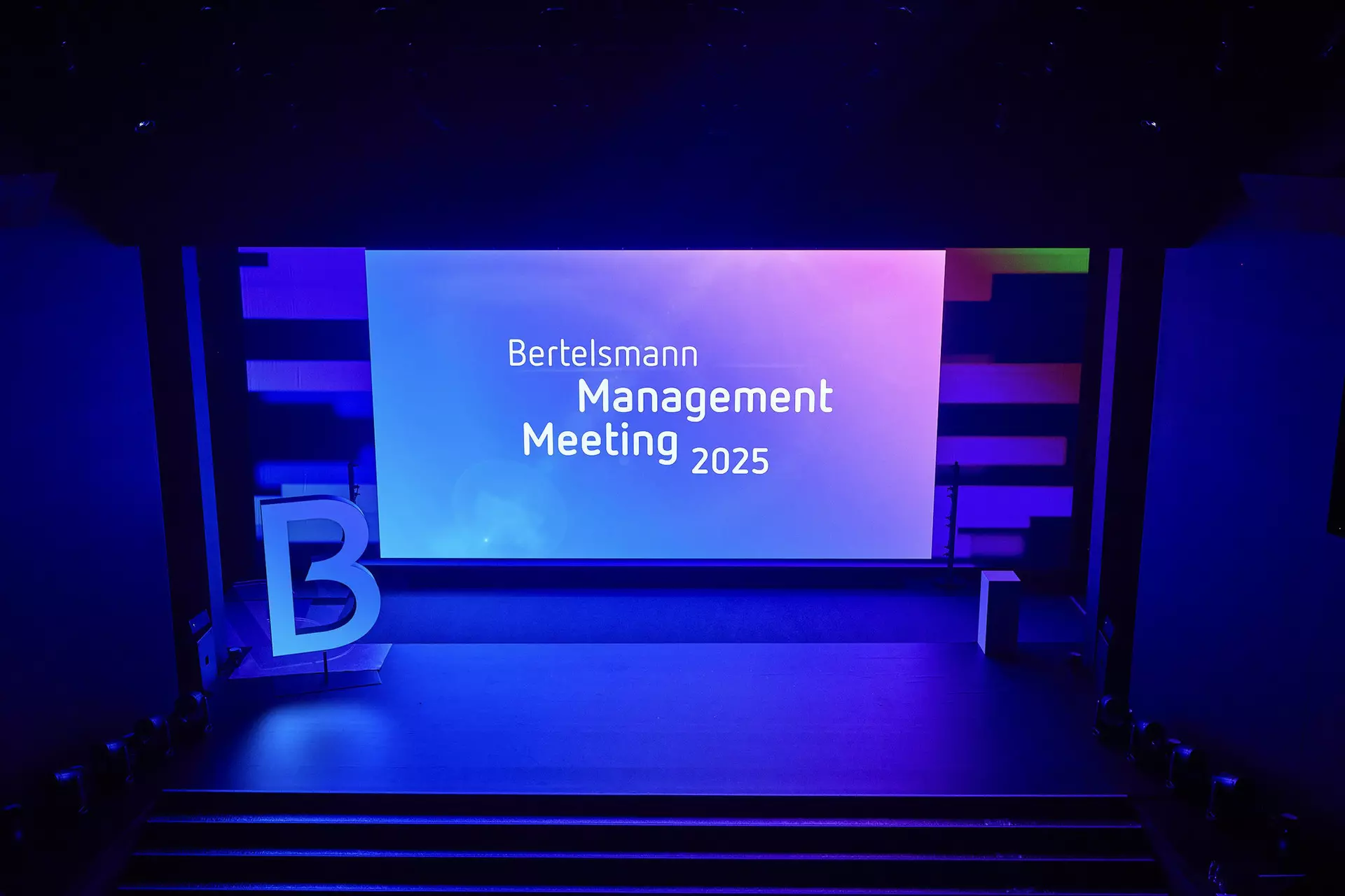 Leere Bühne mit großer Leinwand „Bertelsmann Management Meeting 2025“ und beleuchtetem Buchstaben „B“.
