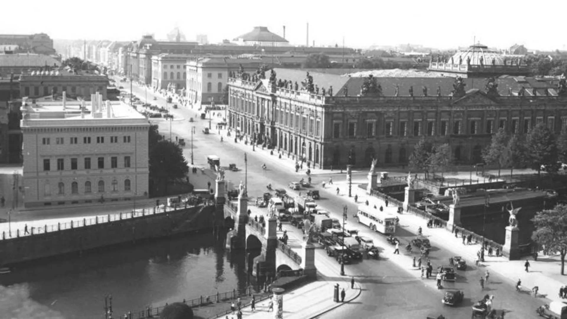 Historische Fotografie mit Blick von oben über eine Brücke mit Statuen, Fluss darunter, belebte Straße, Fahrzeuge, Fußgänger und große mehrgeschossige Gebäude
