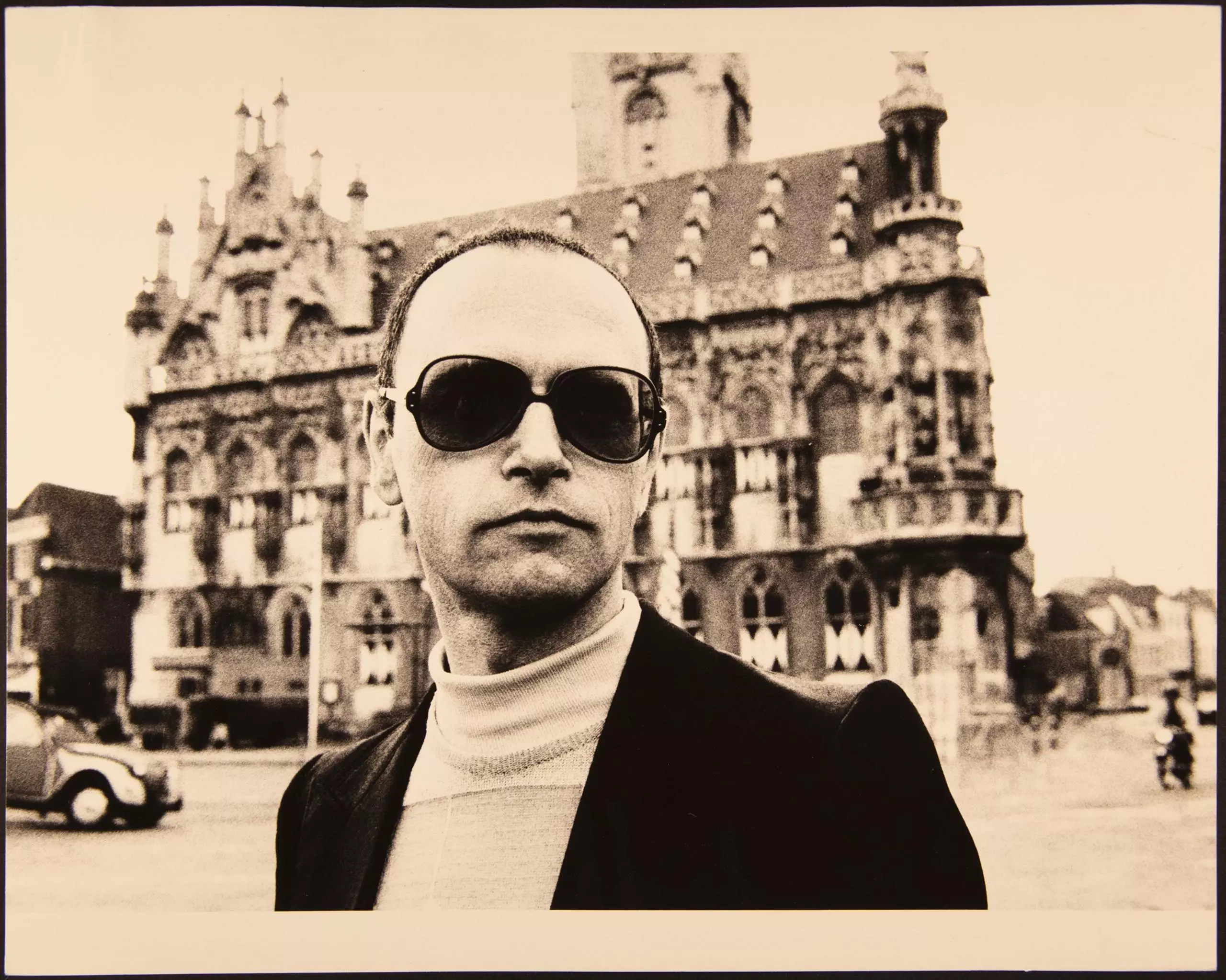 Person mit Sonnenbrille im Anzug vor historischer Gebäudefassade auf Platz.